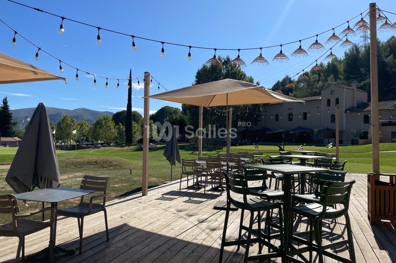 Terrasse en bois avec tables, chaises et parasols, éclairée par des guirlandes lumineuses, donnant sur un espace vert.