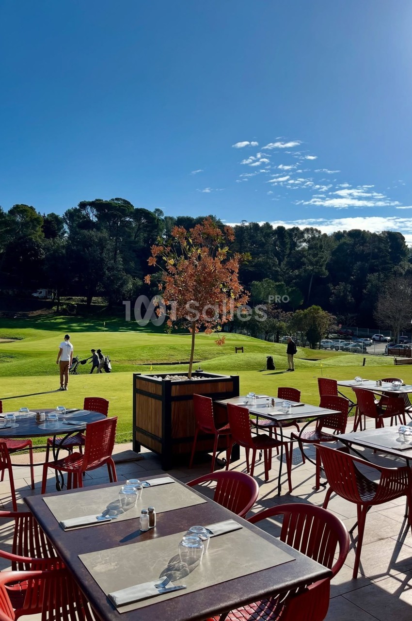 Terrasse d'un restaurant avec des tables vides, vue sur un terrain de golf entouré d'arbres sous un ciel ensoleillé.