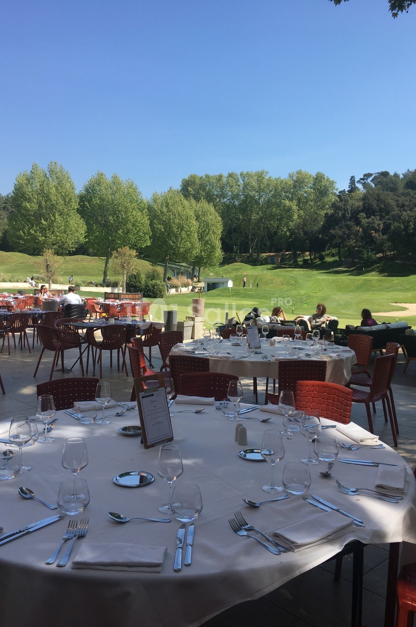 Terrasse d'un restaurant avec des tables dressées, vue sur un terrain de golf entouré d'arbres sous un ciel bleu.