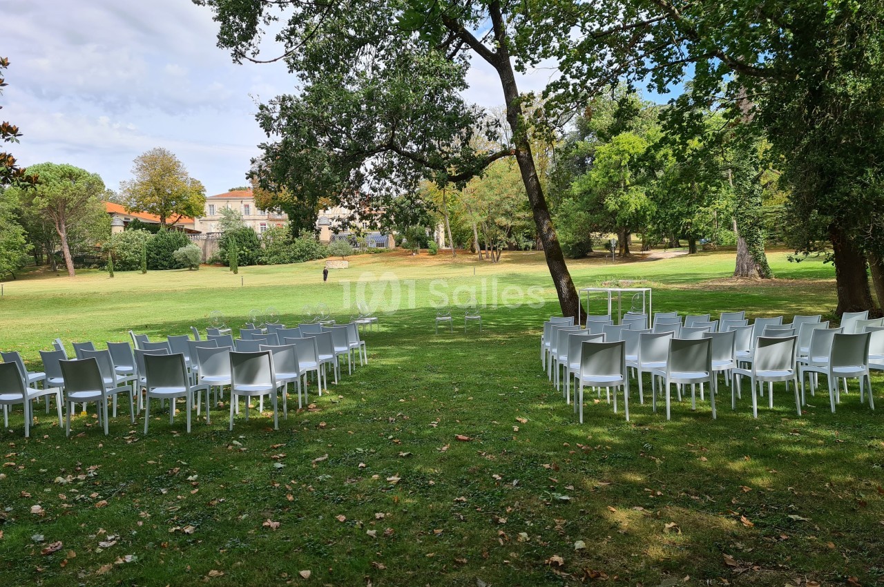 Chaises blanches disposées en extérieur sur une pelouse, entourées d'arbres, avec une arche au fond pour une cérémonie.