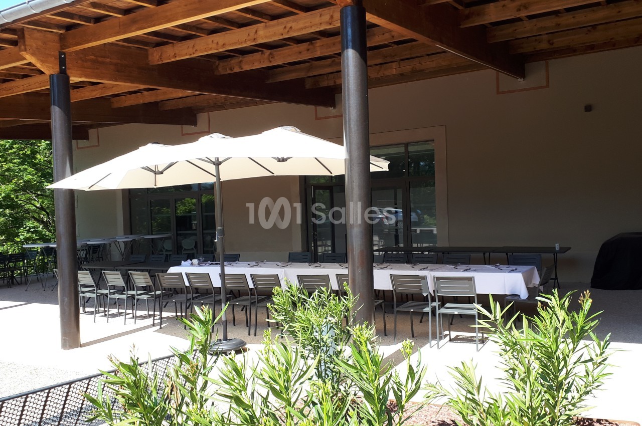 Terrasse ombragée avec une grande table dressée sous un parasol, entourée de chaises et de végétation.