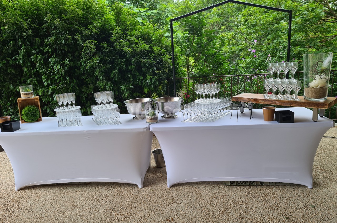Tables décorées avec nappes blanches, verres, seaux à glace et éléments naturels, disposées en extérieur verdoyant.