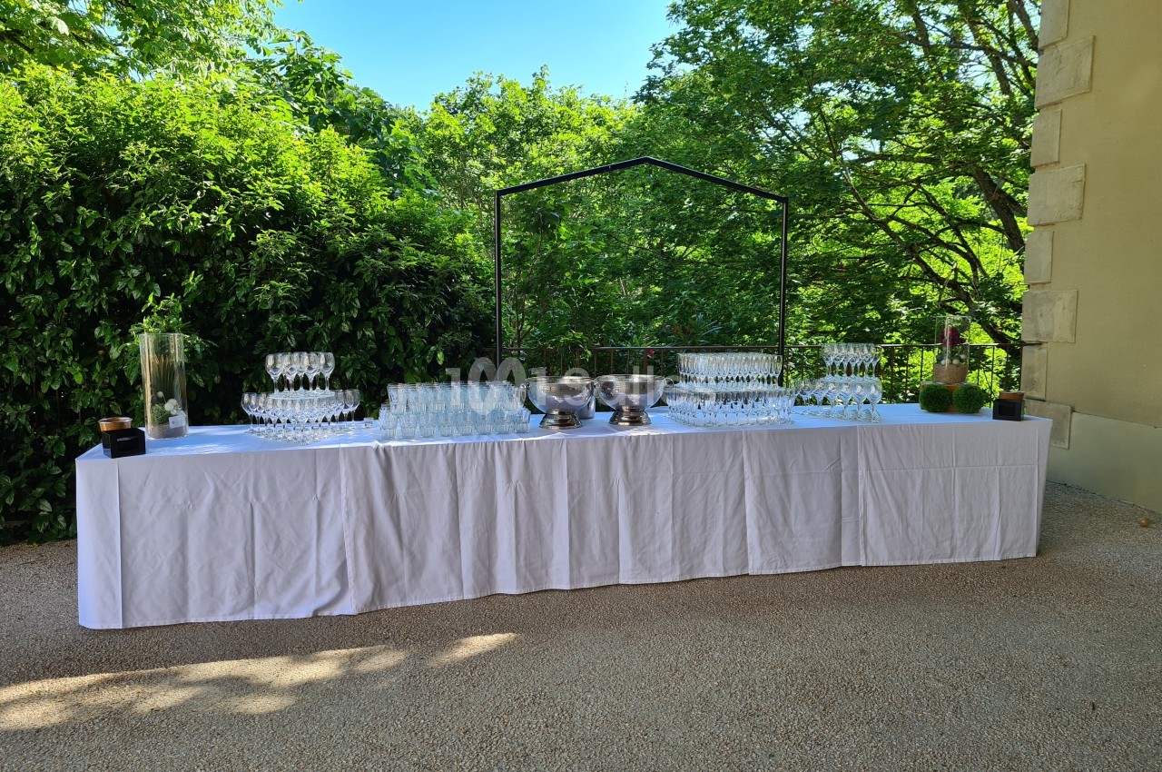 Table dressée en extérieur avec nappage blanc, verres à pied, seaux à champagne et verdure en arrière-plan.