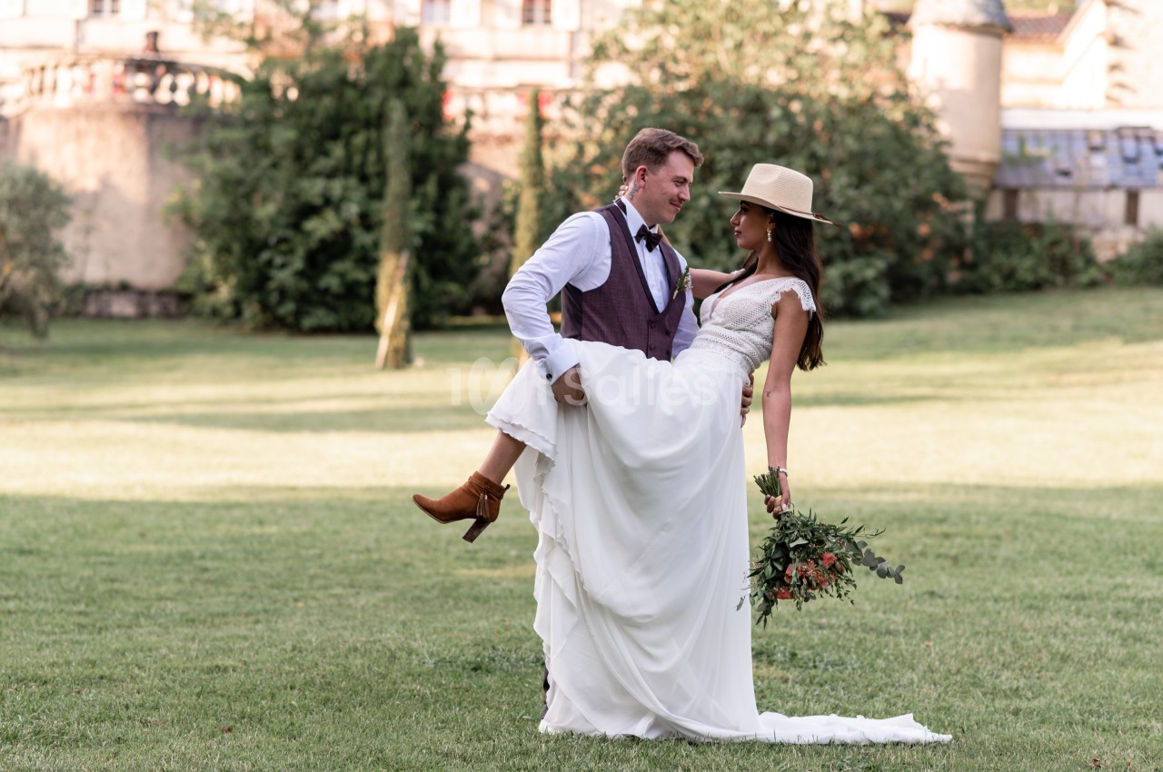 Un homme en costume porte une femme en robe de mariée tenant un bouquet, dans un parc verdoyant près d'un bâtiment ancien.