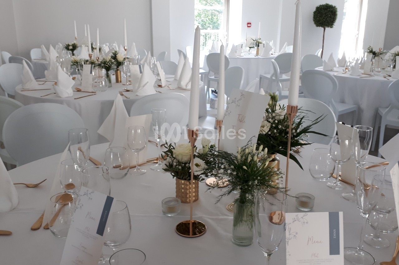 Salle de réception décorée avec des tables rondes, nappes blanches, bougies, fleurs et serviettes pliées.