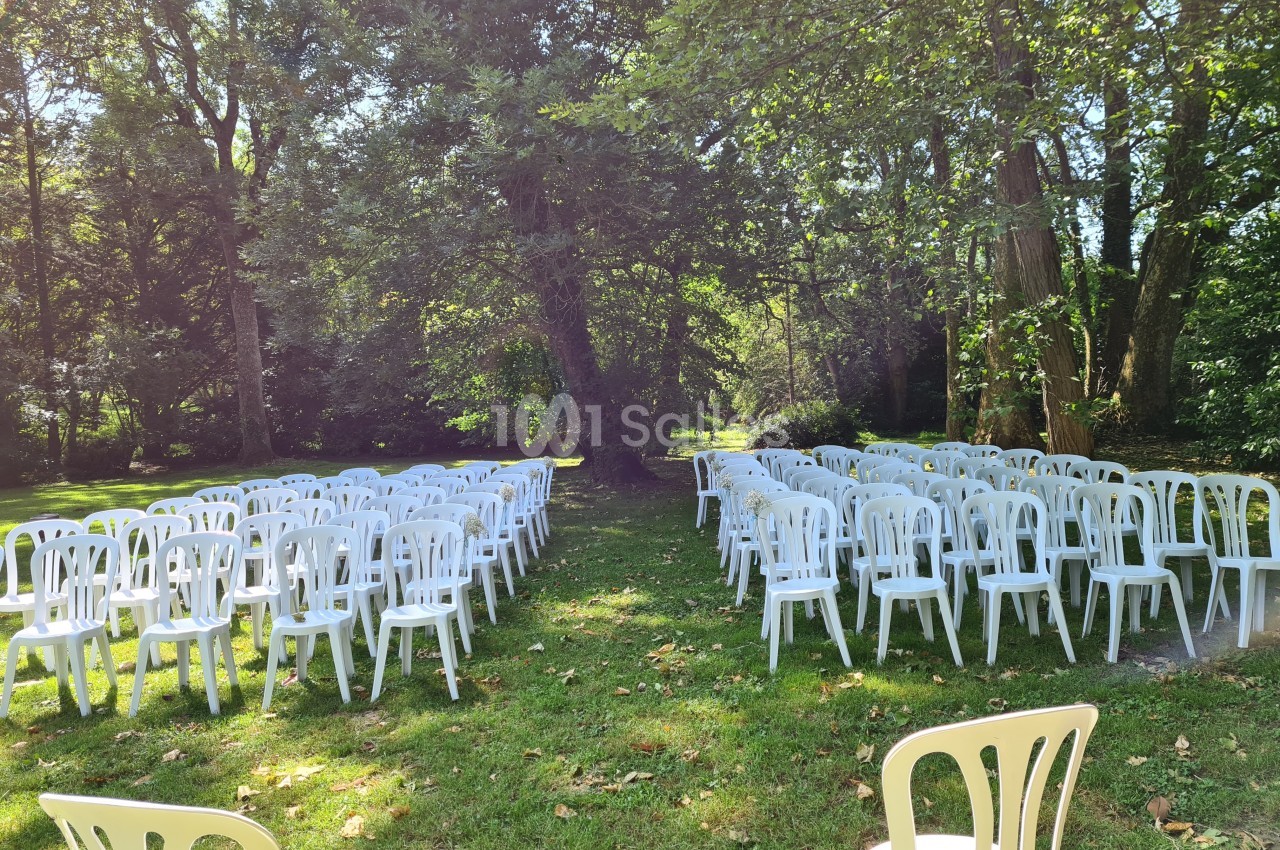 Chaises blanches disposées en rangées sur une pelouse, entourées d'arbres dans un cadre extérieur ensoleillé.