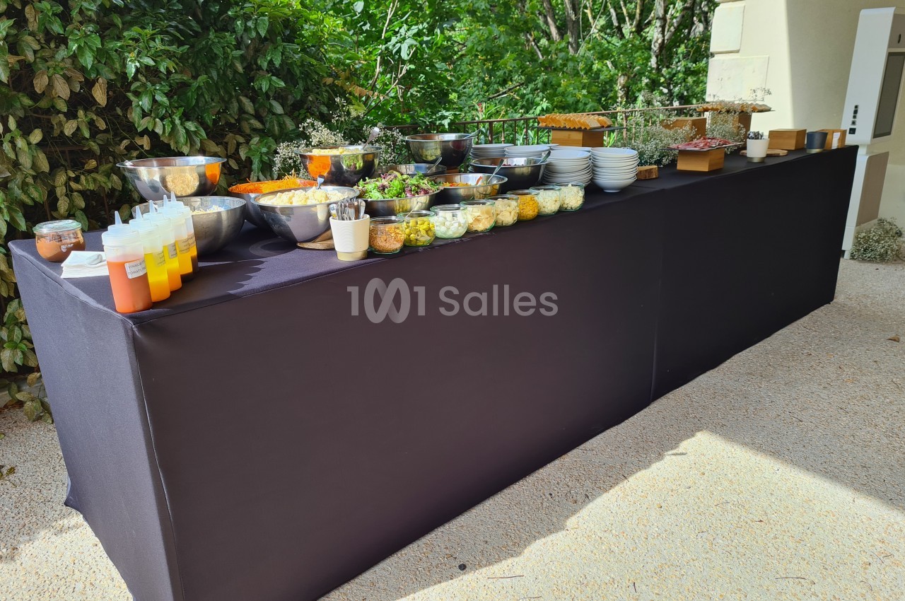 Buffet en plein air avec salades, sauces et plats disposés sur une table noire, entourée de verdure.