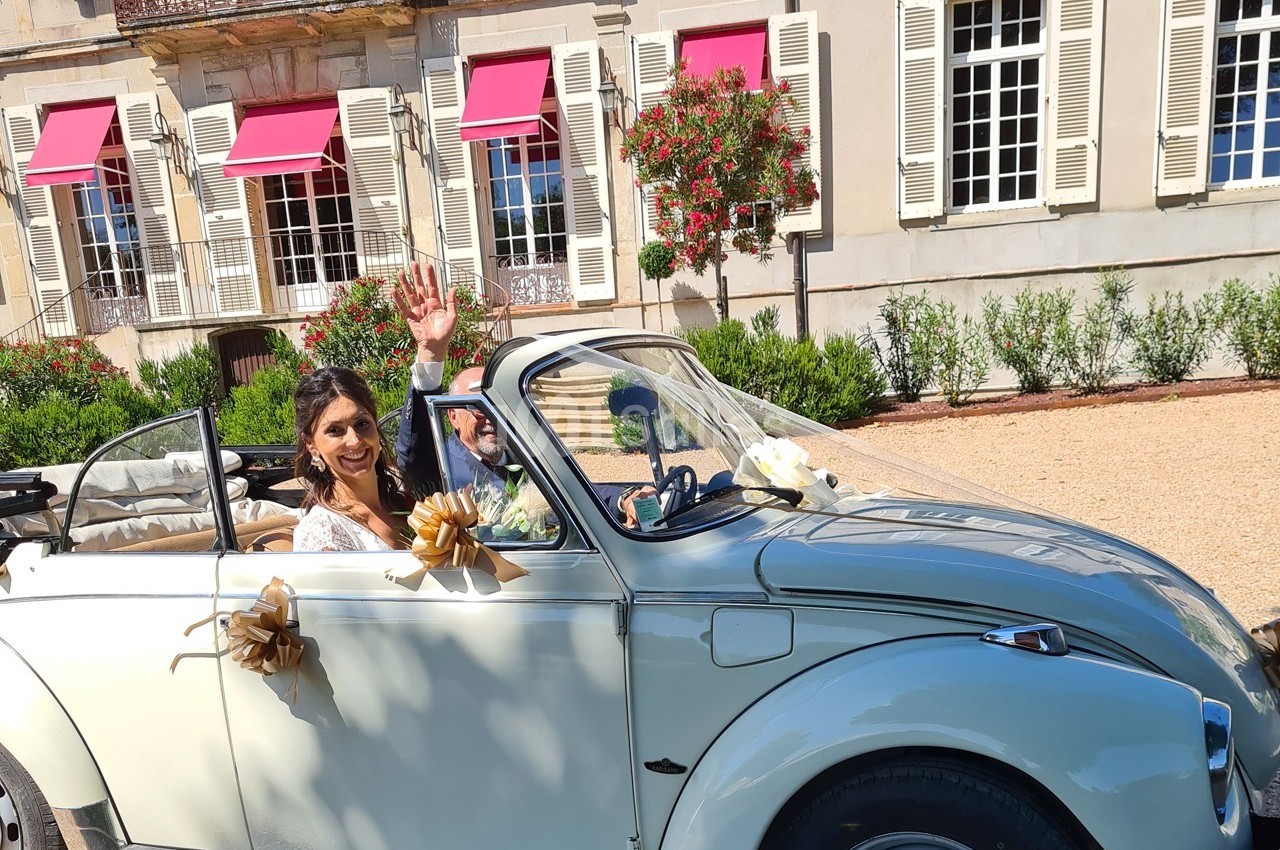 Une mariée souriante salue depuis une voiture décapotable décorée, devant un bâtiment avec volets blancs et stores rouges.