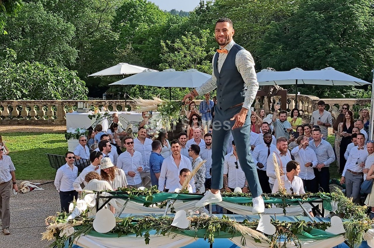 Un homme en costume se tient debout sur une table décorée lors d'un événement en plein air avec des invités en arrière-plan.
