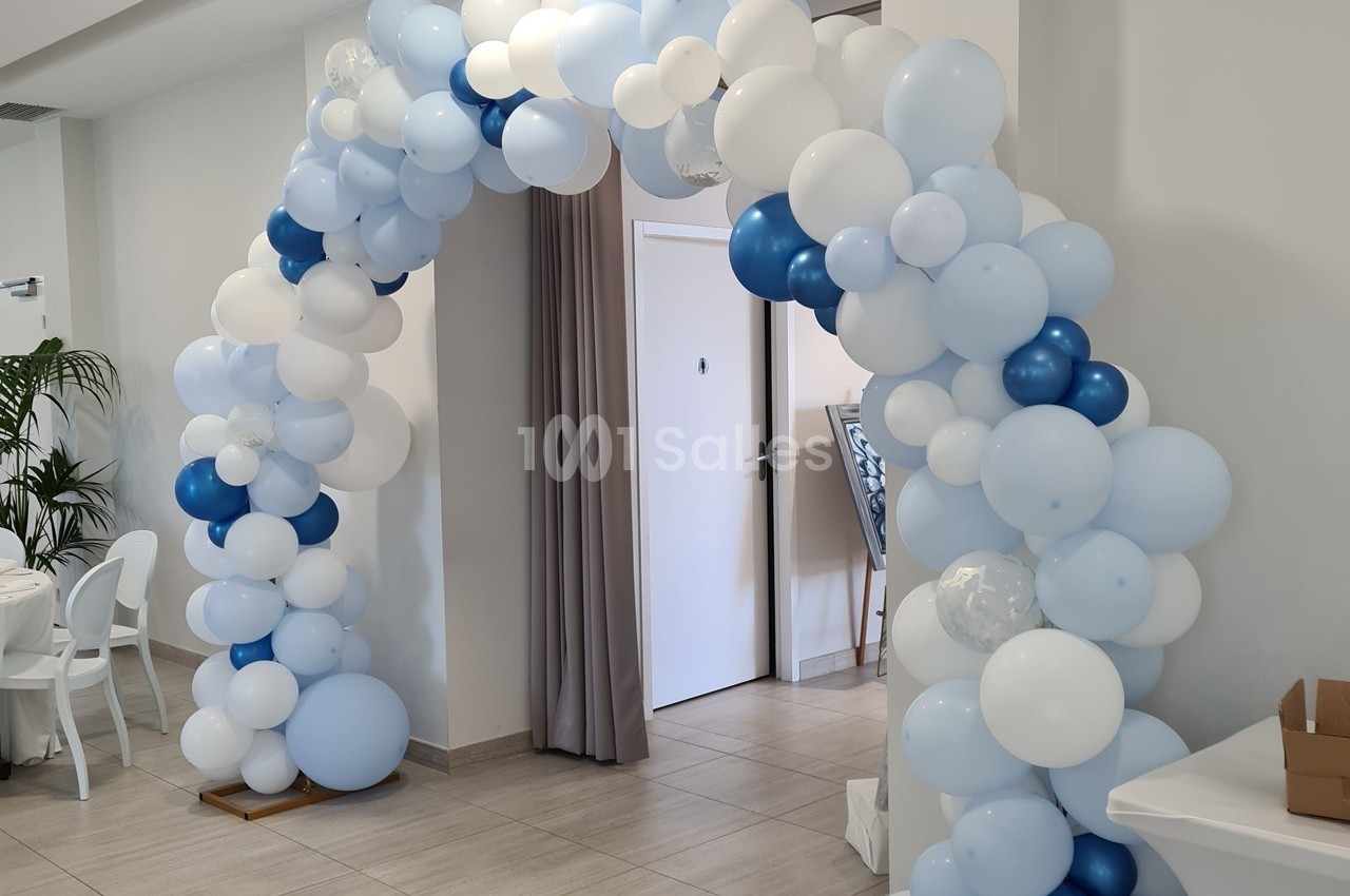 Arche décorative en ballons blancs et bleus installée dans une salle lumineuse avec des murs clairs.