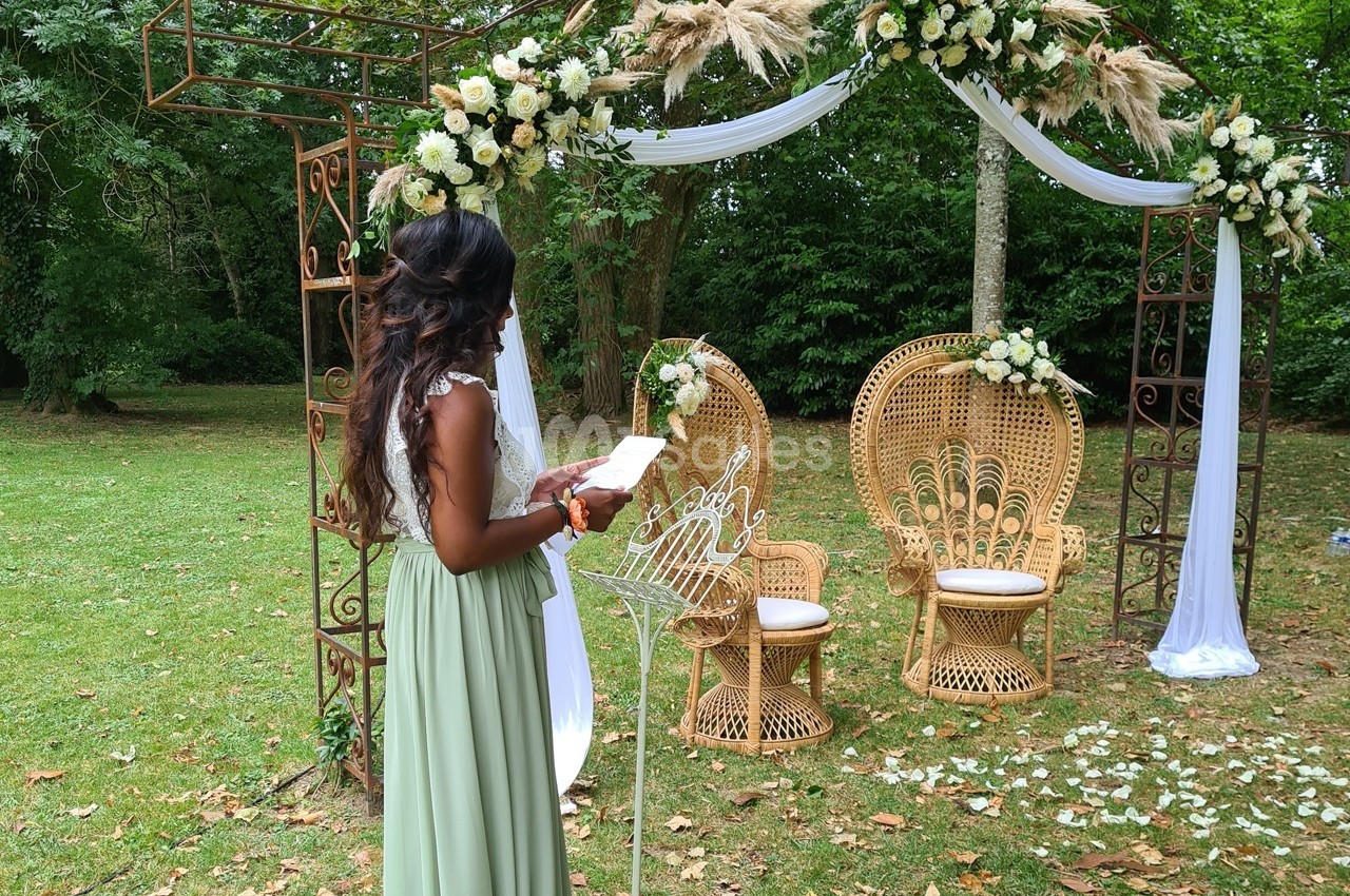 Une femme en robe verte se tient devant une arche décorée de fleurs et de chaises en osier dans un jardin.