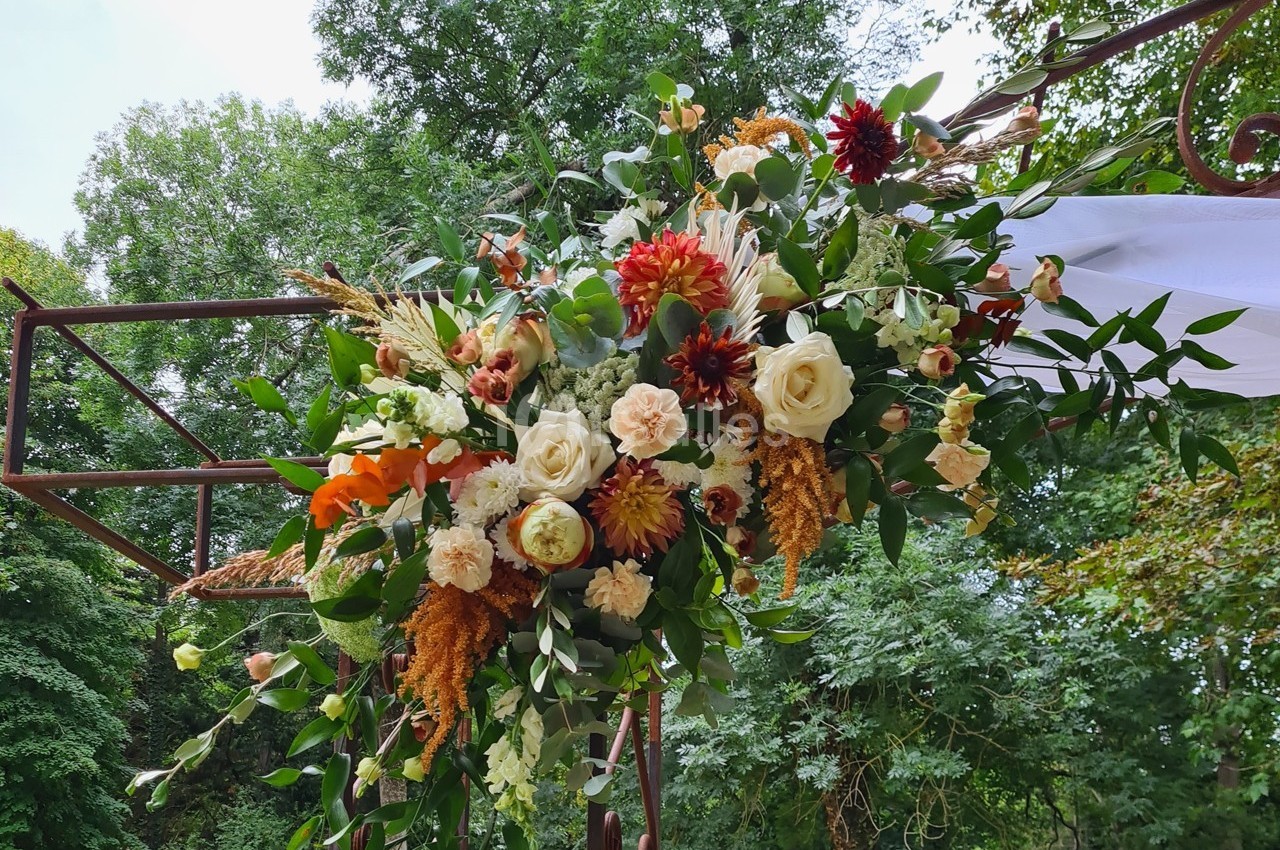 Arrangement floral coloré avec roses, dahlias et feuillage, suspendu à une structure métallique en extérieur verdoyant.