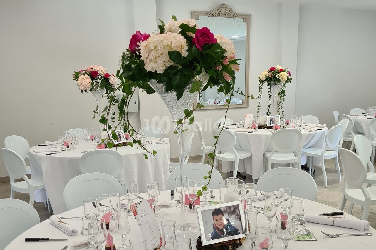 Salle de réception décorée avec des tables rondes, nappes blanches, centres de table fleuris et photo encadrée.