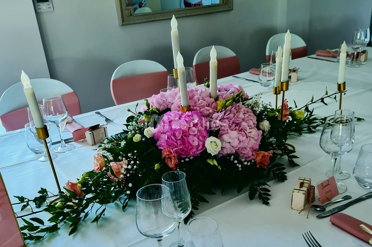 Centre de table floral avec hortensias roses, bougies blanches et vaisselle disposée sur une nappe blanche.