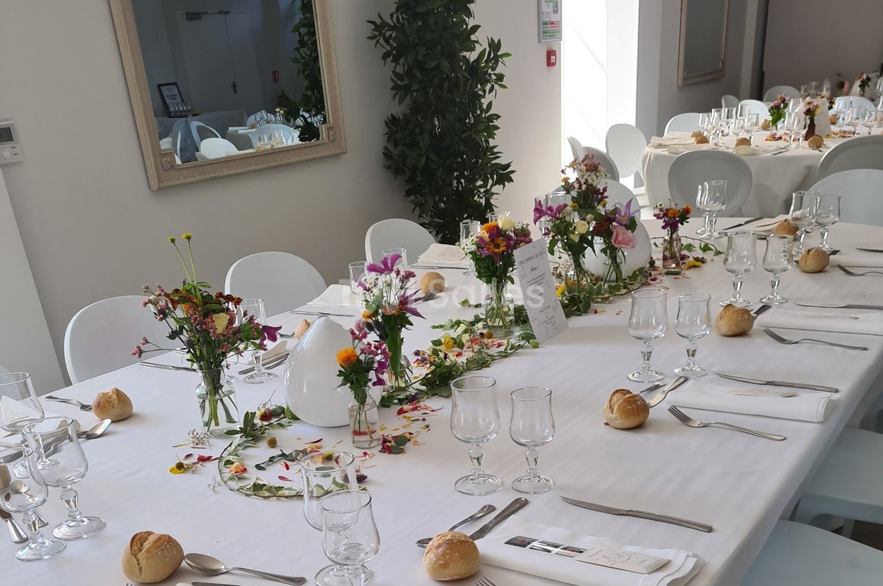 Table dressée pour un repas festif avec nappes blanches, fleurs colorées, verres, couverts et petits pains.