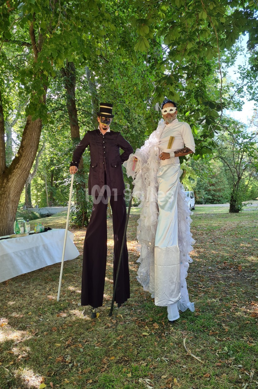 Deux personnes sur échasses, vêtues de costumes élaborés et masques, posent dans un parc verdoyant.