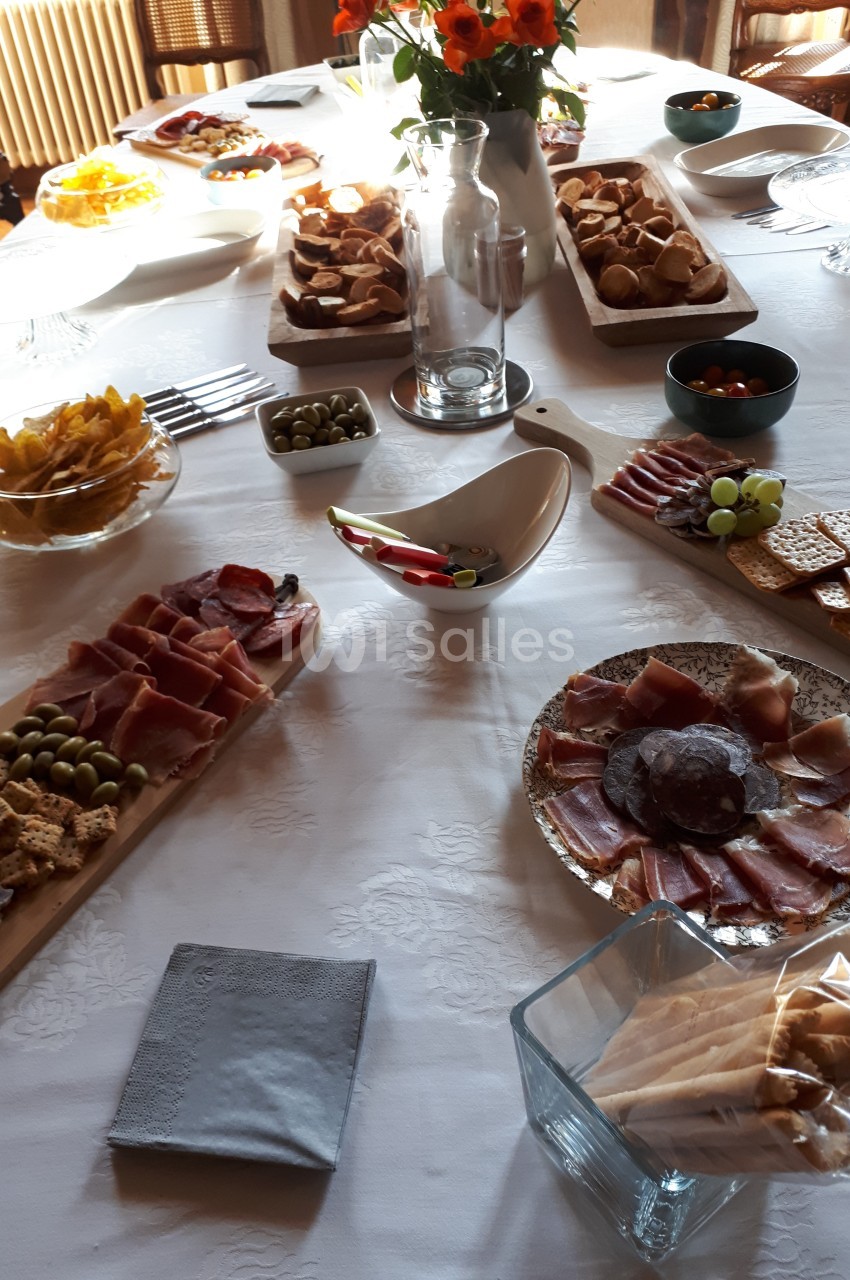 Table dressée avec des plateaux de charcuterie, fromages, olives, biscuits apéritifs et un bouquet de fleurs au centre.