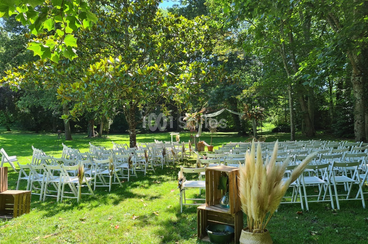 Chaises blanches disposées en extérieur pour une cérémonie, entourées d'arbres et décorées de fleurs et de pampas.