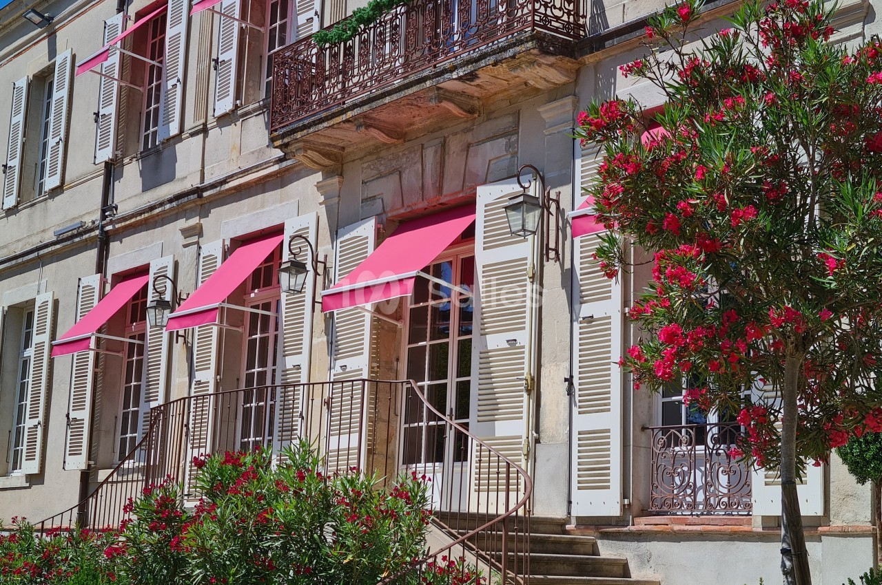 Façade d'une maison en pierre avec volets blancs, stores roses et un jardin fleuri au premier plan.