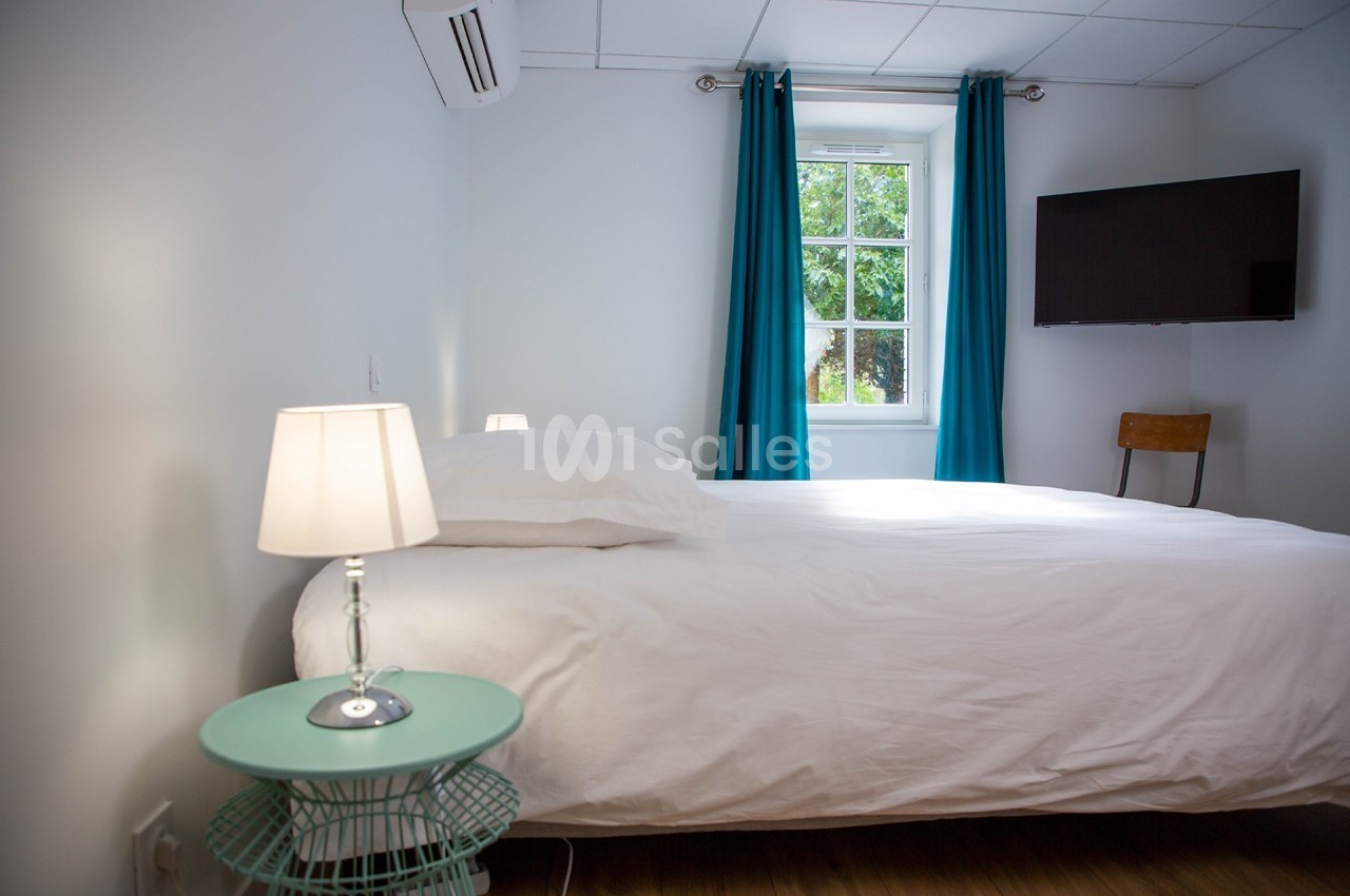 Chambre lumineuse avec lit double, table de chevet, lampe, fenêtre avec rideaux bleus et télévision fixée au mur.