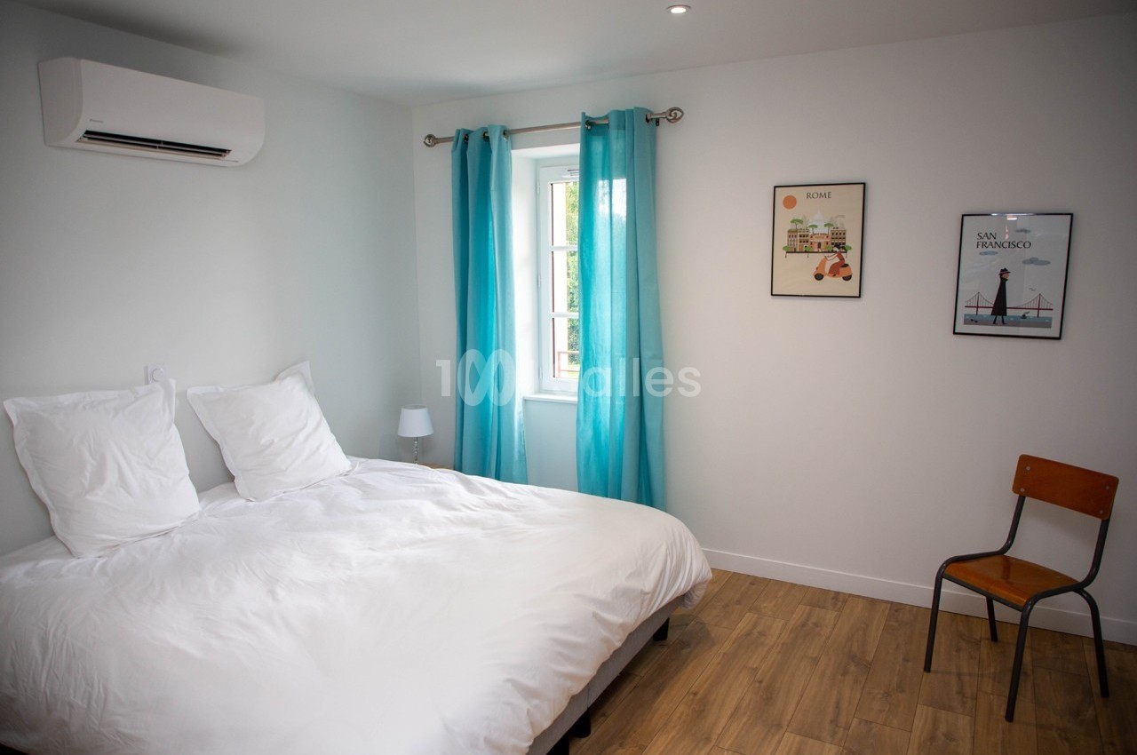 Chambre lumineuse avec lit double, rideaux bleus, chaise en bois, affiches murales et sol en parquet clair.