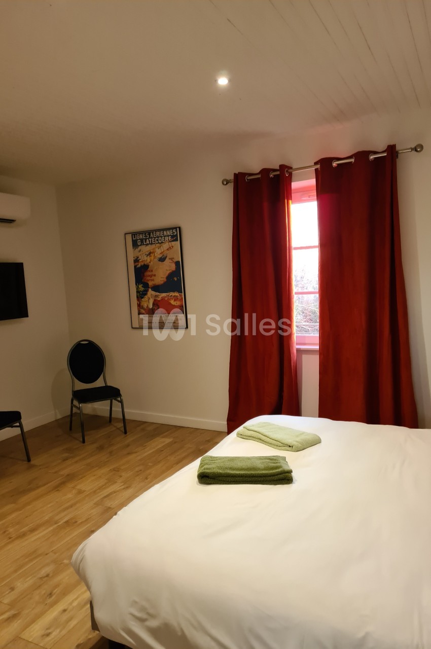 Chambre lumineuse avec lit double, draps blancs, serviettes vertes, rideaux rouges et télévision murale.