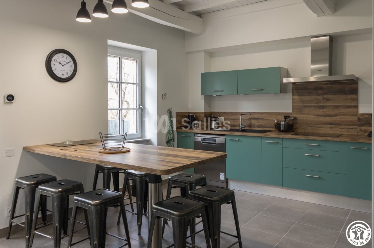 Cuisine moderne avec meubles verts, plan de travail en bois, tabourets métalliques et éclairage suspendu.
