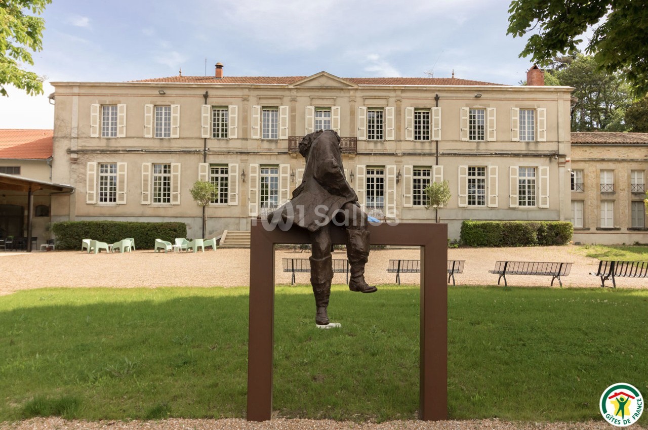 Statue d'un homme assis sur un cadre métallique devant un bâtiment ancien avec des fenêtres symétriques.
