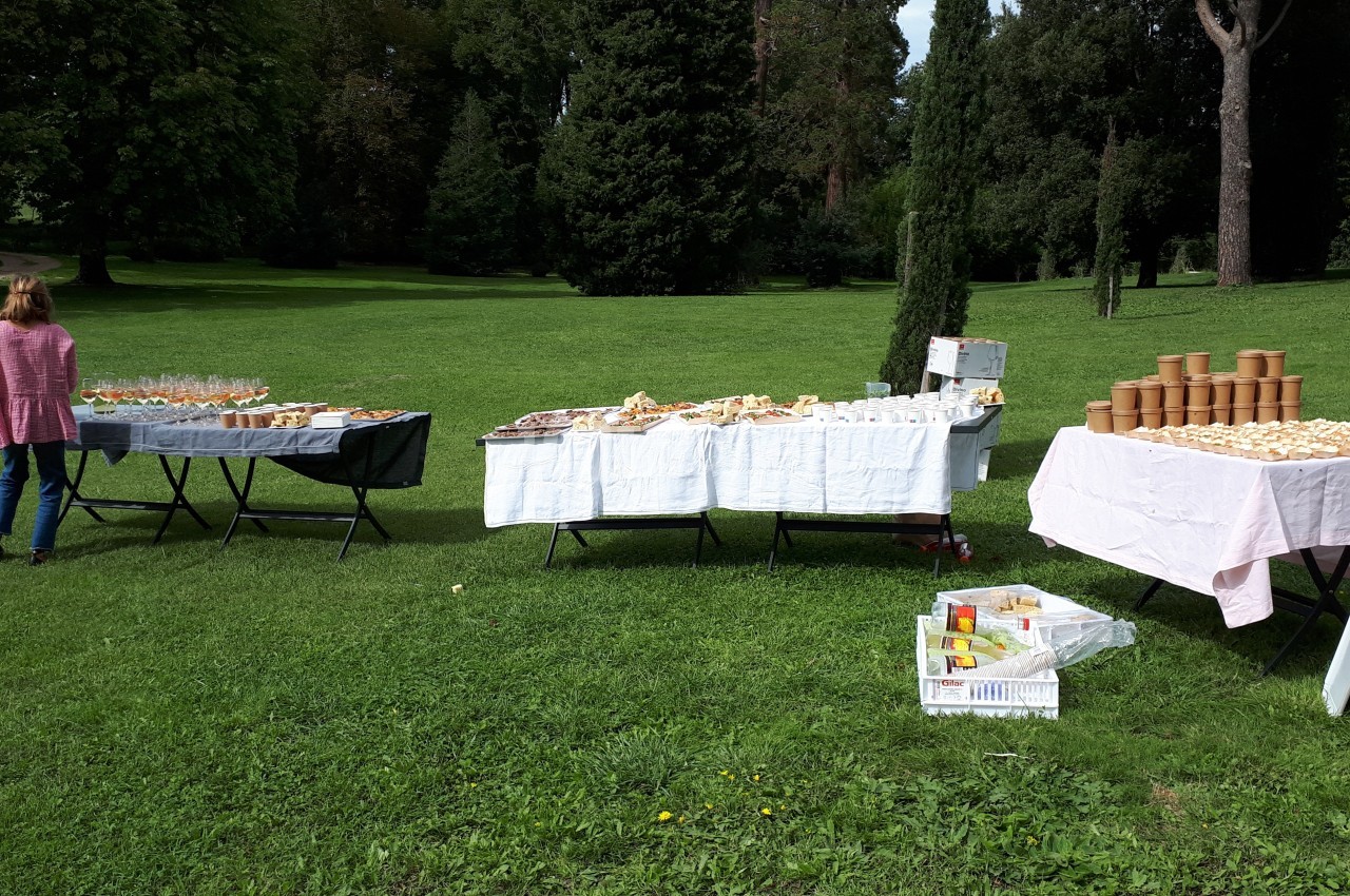 Tables dressées en extérieur avec nappes blanches, contenant des aliments, boissons et pots, dans un parc verdoyant.