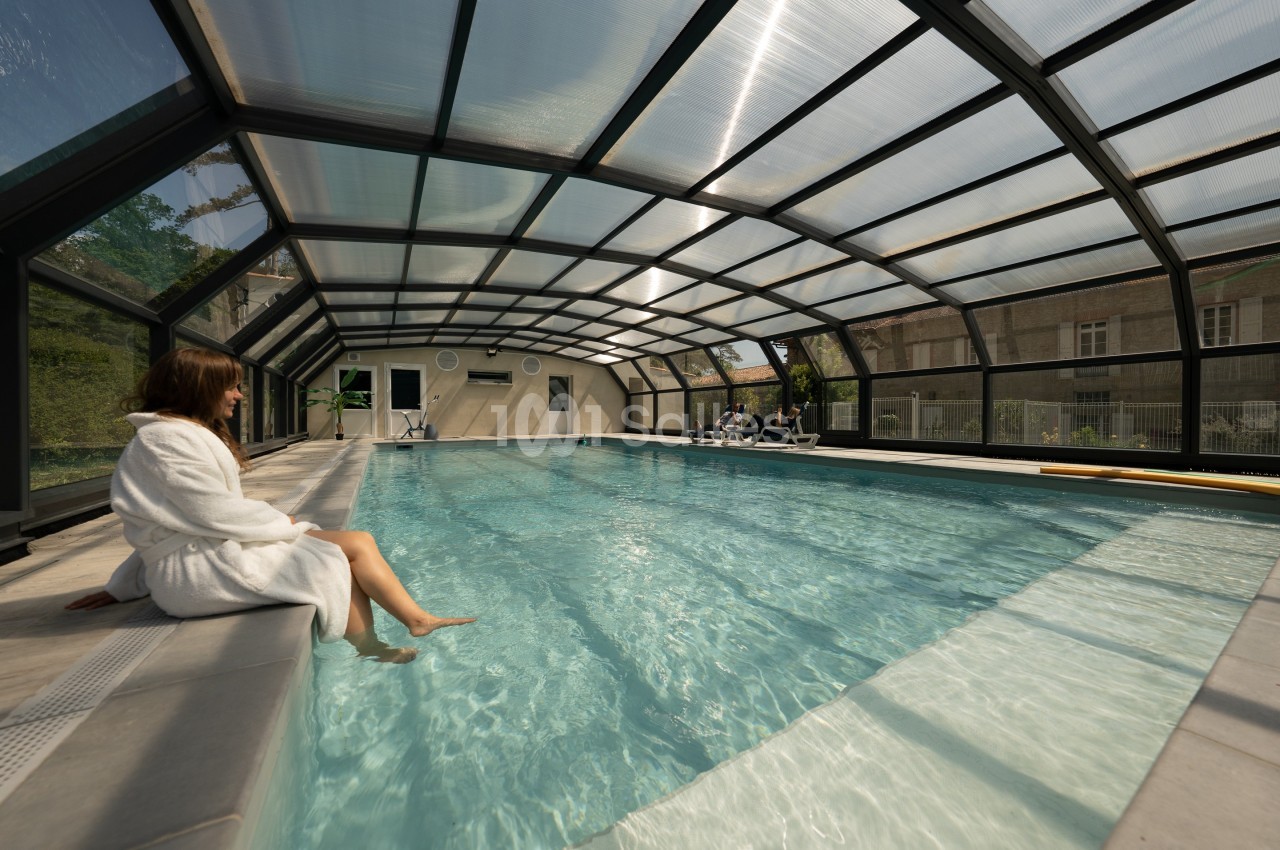 Femme en peignoir assise au bord d'une piscine couverte, avec des baigneurs en arrière-plan.