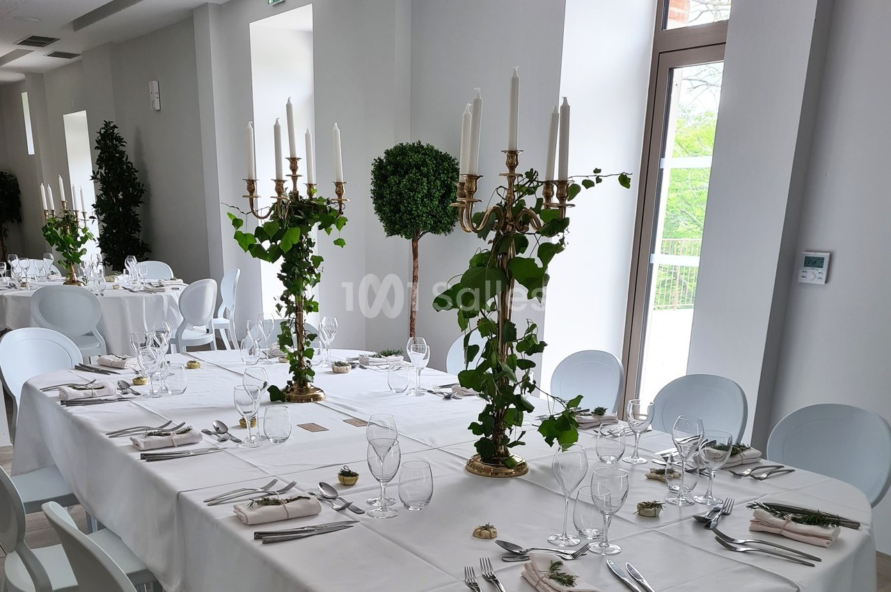 Salle de réception lumineuse avec tables dressées, chandeliers ornés de feuillage et grandes fenêtres donnant sur l…