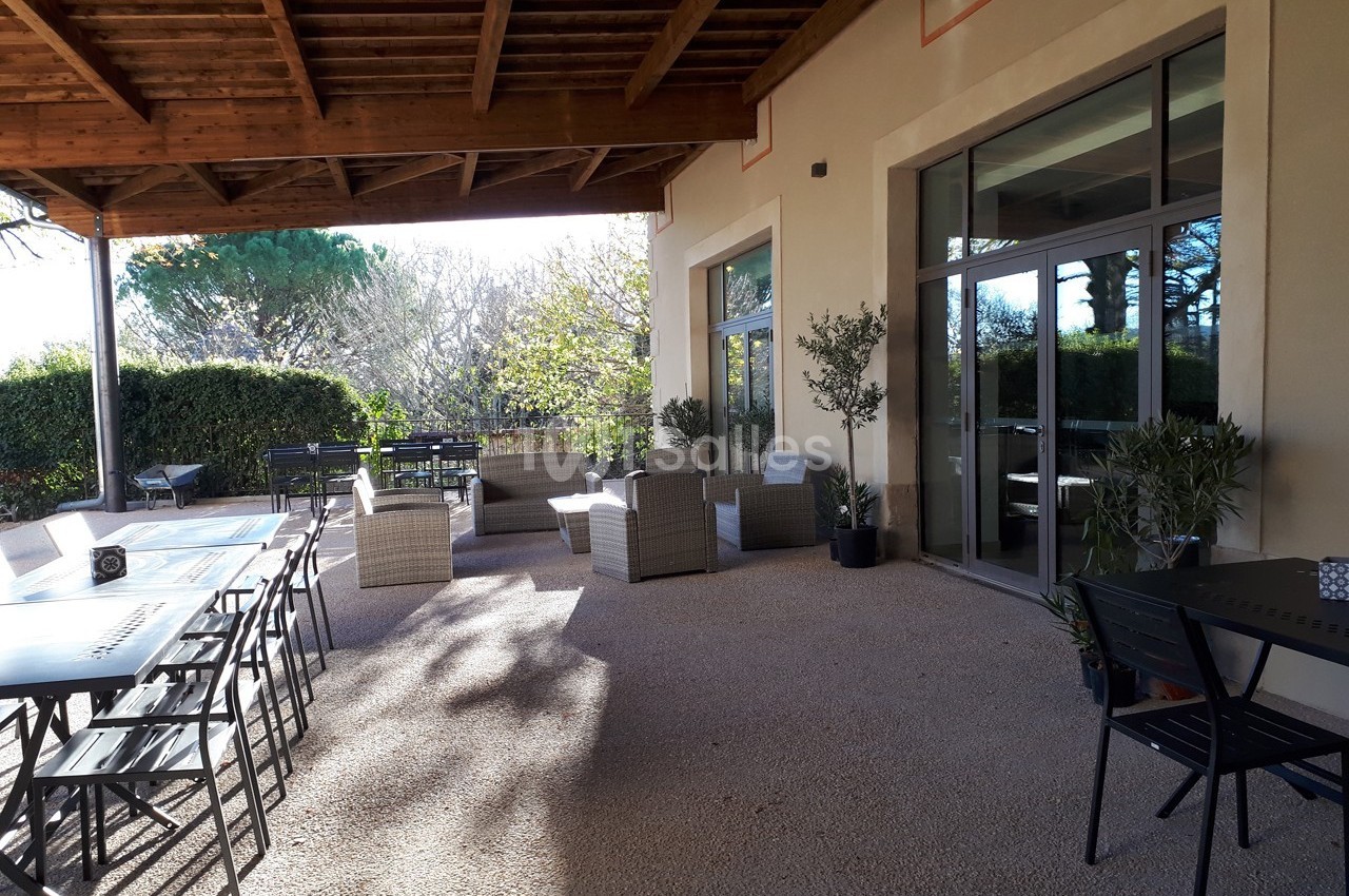 Terrasse extérieure avec tables, chaises et fauteuils sous une pergola, entourée de verdure et baignée de lumière naturelle.