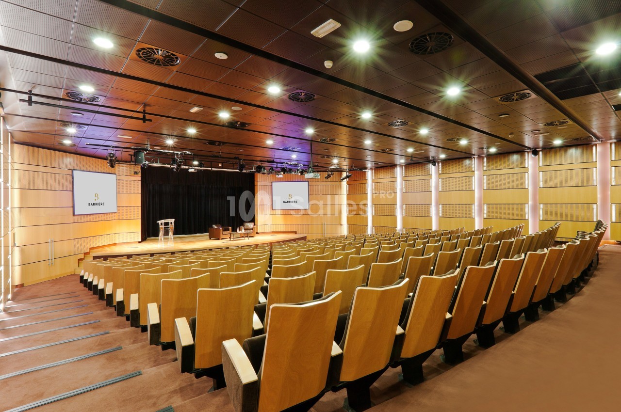 Salle de conférence moderne avec rangées de sièges en bois, scène équipée et écrans affichant un logo.