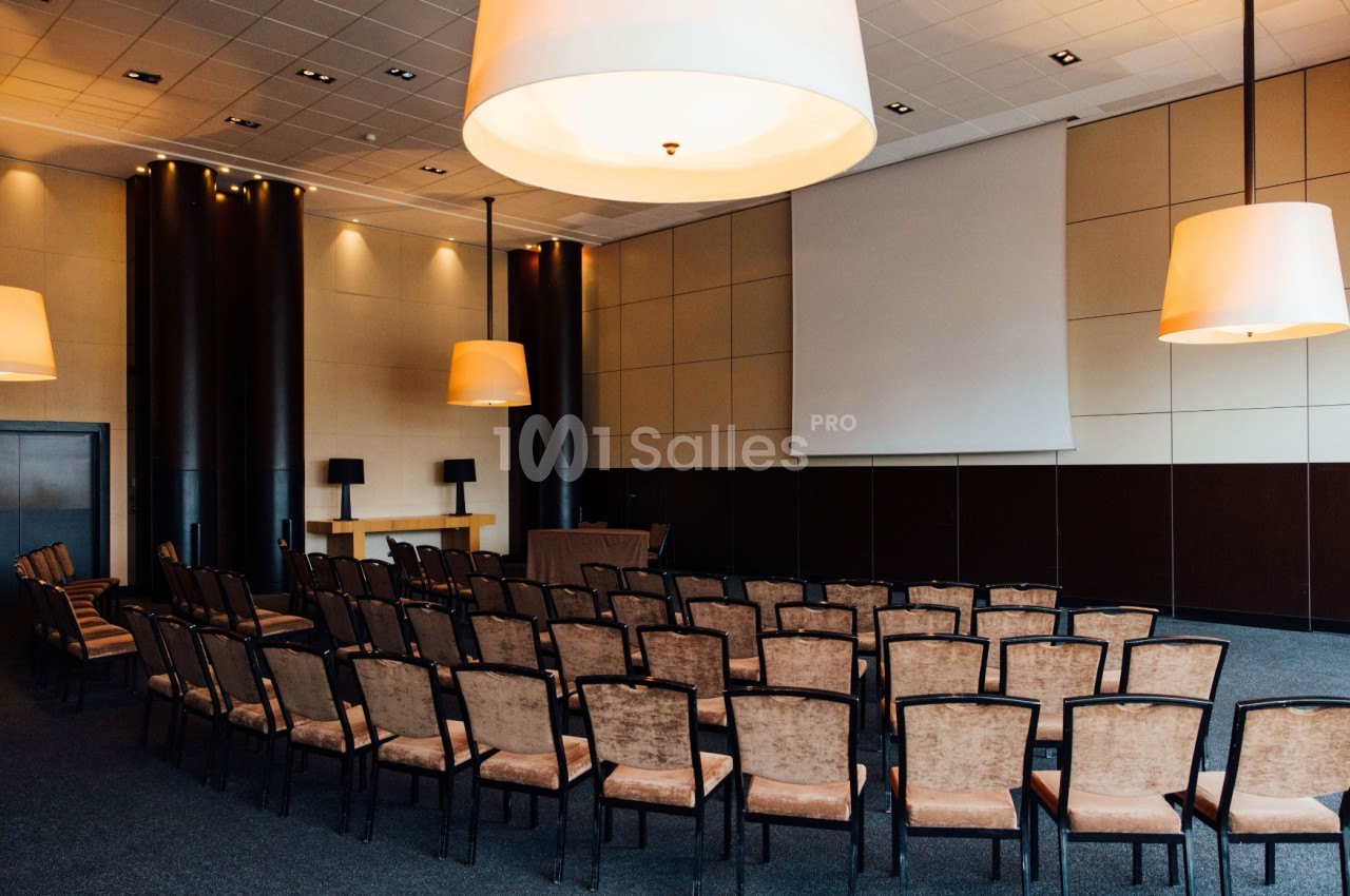 Salle de conférence vide avec des rangées de chaises alignées face à un écran de projection et un pupitre.