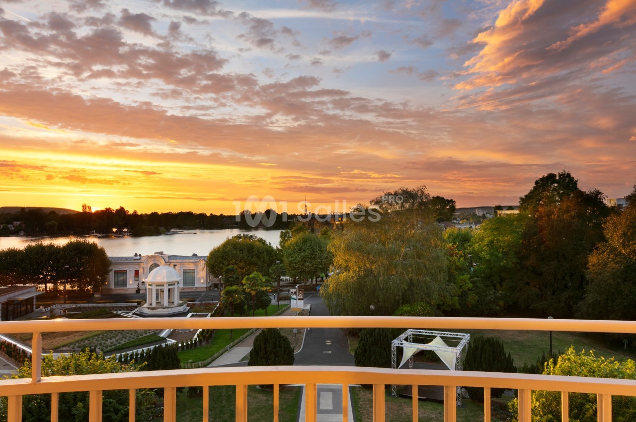 Vue d'un parc avec un pavillon, des arbres et un lac au coucher du soleil, depuis une balustrade jaune.