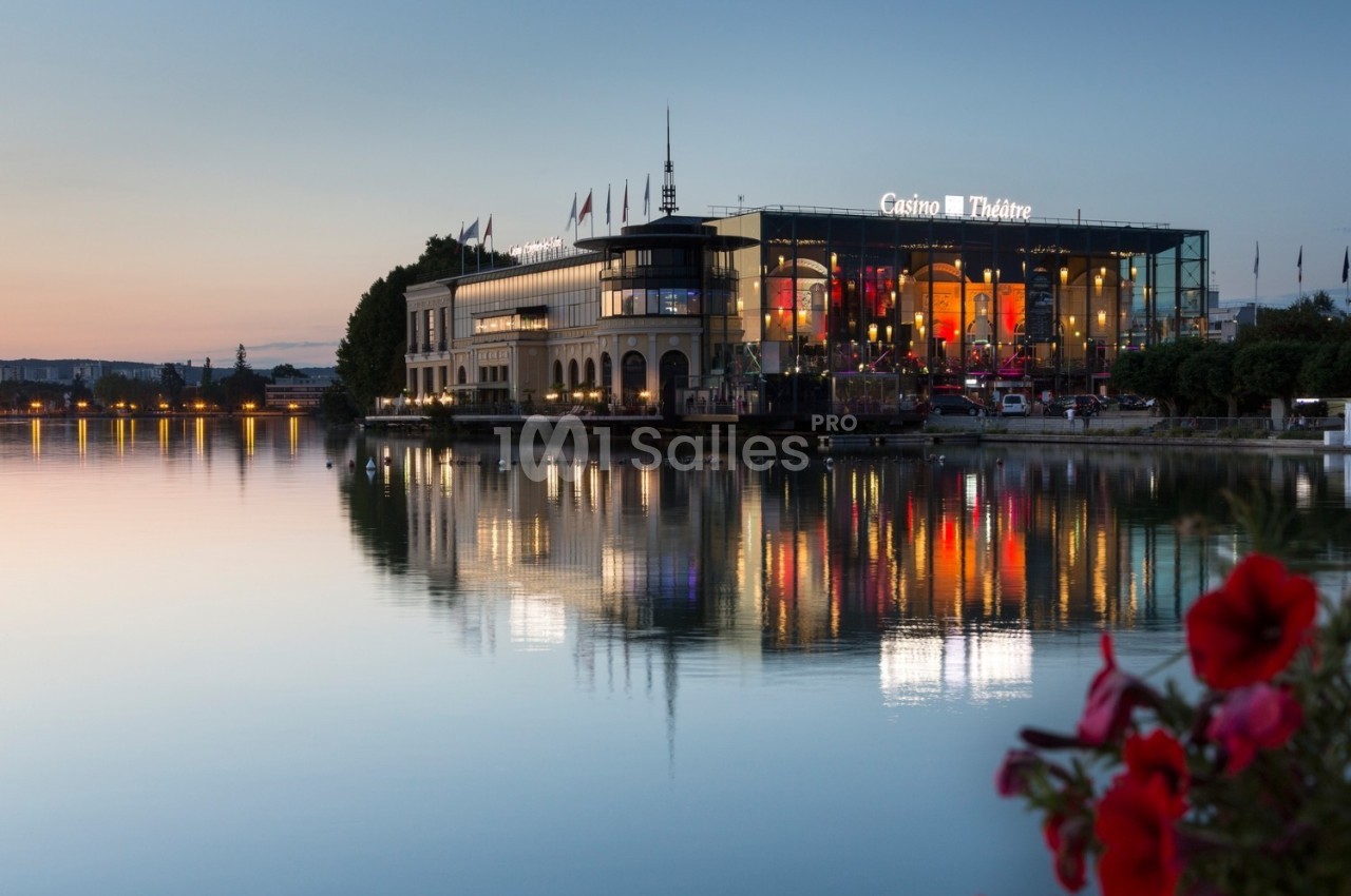Bâtiment d'un casino-théâtre illuminé au bord d'un lac au crépuscule, avec des reflets sur l'eau calme.
