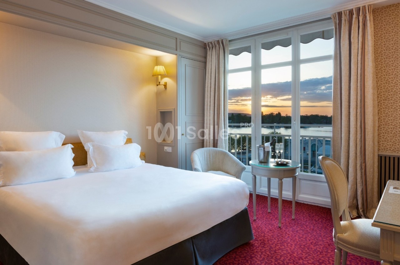 Chambre lumineuse avec lit double, fauteuils, table et grande fenêtre offrant une vue sur un coucher de soleil.