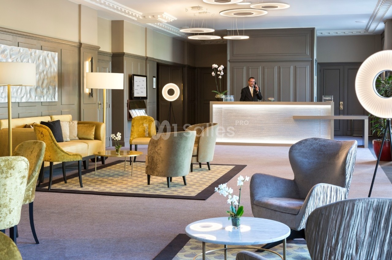 Hall d'hôtel moderne avec fauteuils, tables basses, éclairage design et réceptionniste au comptoir lumineux.