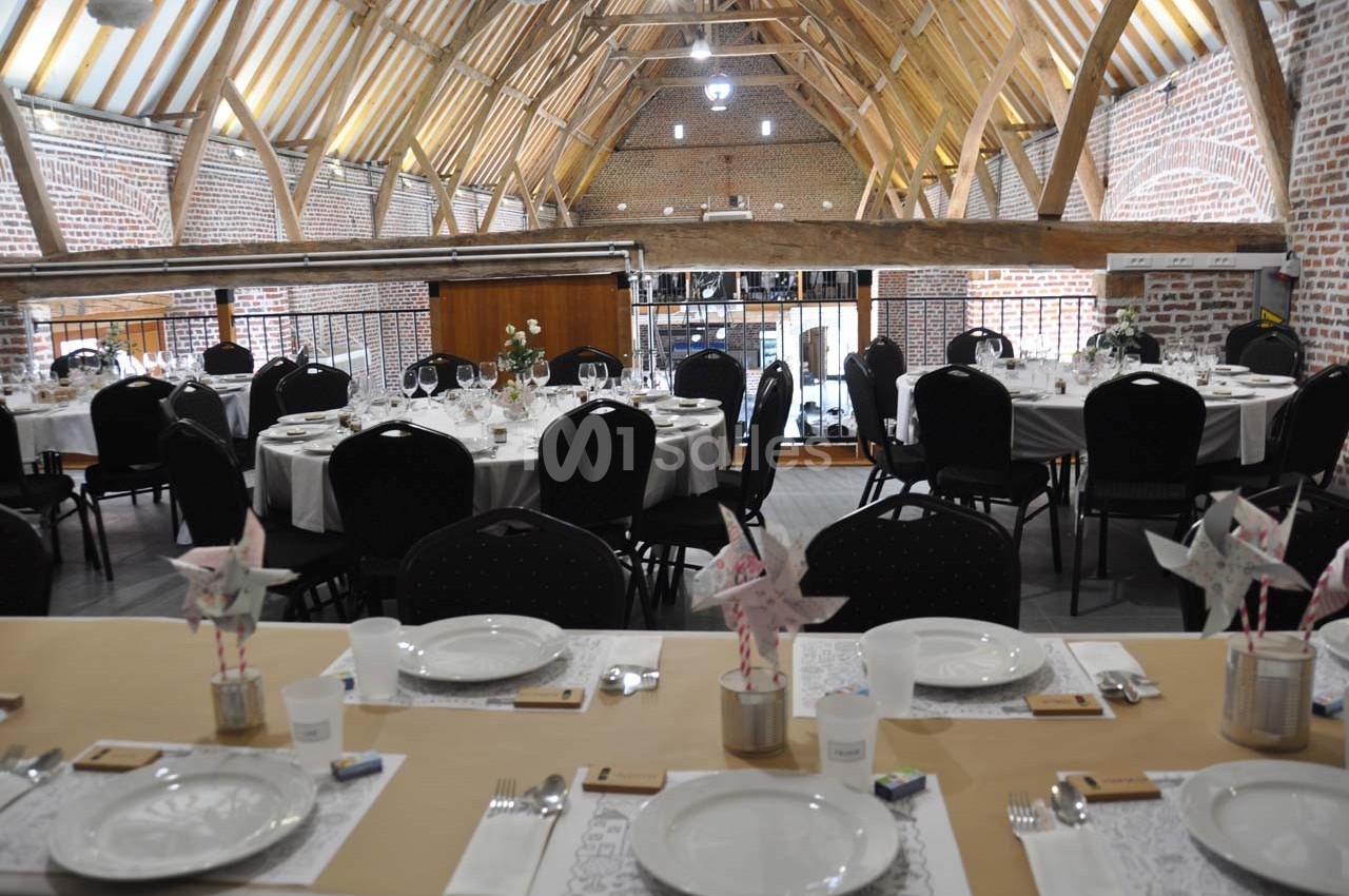 Salle de réception dans une grange rénovée avec tables dressées pour un événement, décorée de moulins à vent en papier.
