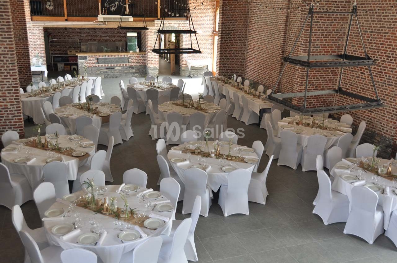 Salle de réception avec tables rondes dressées, nappes blanches et décoration sobre, dans un espace en briques apparentes.