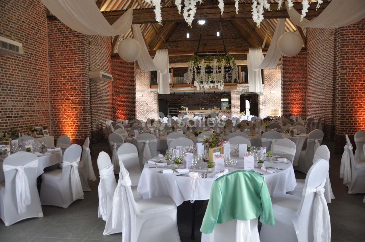 Salle de réception avec tables décorées, nappes blanches, chaises couvertes et guirlandes suspendues sous un plafond en bois.