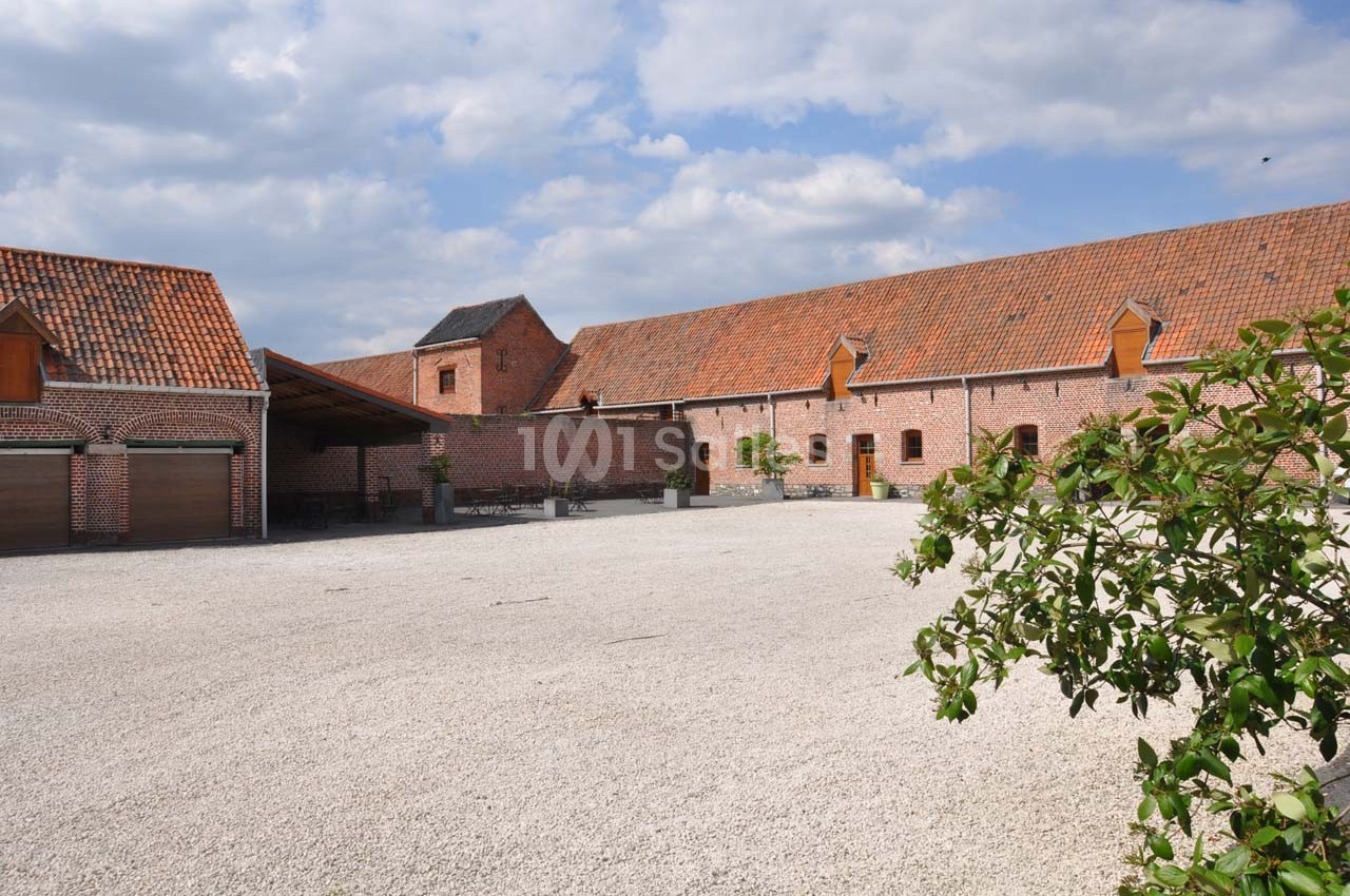 Cour pavée entourée de bâtiments en briques rouges avec toits en tuiles, sous un ciel partiellement nuageux.