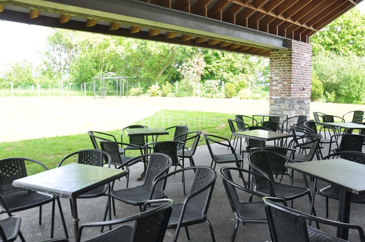 Terrasse couverte avec des tables et chaises noires, donnant sur un jardin verdoyant par une journée ensoleillée.