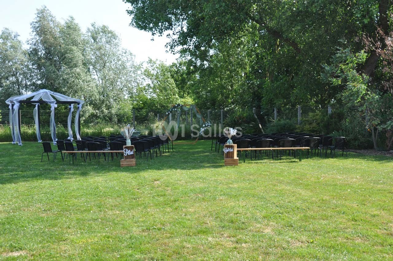 Chaises alignées en extérieur devant une arche décorée pour une cérémonie, entourées de verdure et d'arbres.
