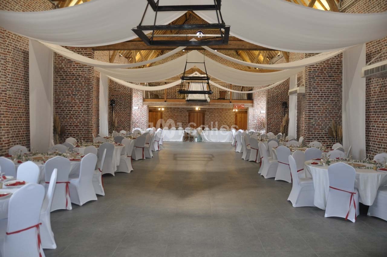 Salle de réception spacieuse avec murs en briques, tables rondes dressées et plafond décoré de voilages blancs.
