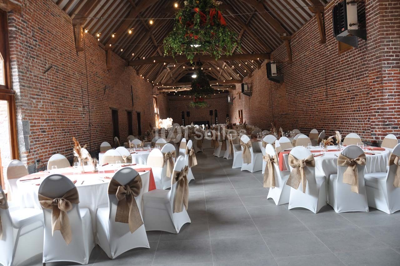 Salle de réception en briques avec tables rondes dressées, chaises décorées de nœuds en toile de jute et guirlandes…