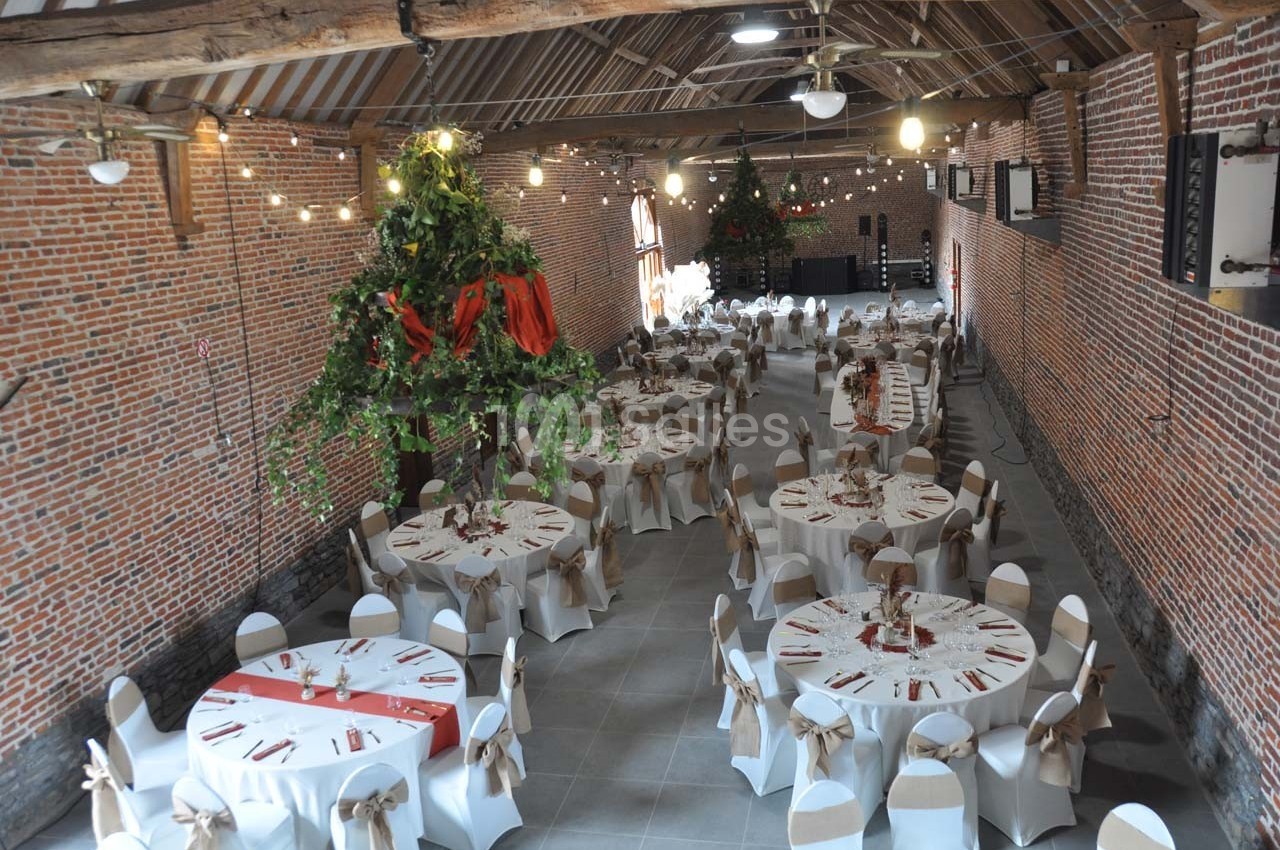 Salle de réception en briques avec tables rondes décorées, chaises blanches et guirlandes lumineuses suspendues.