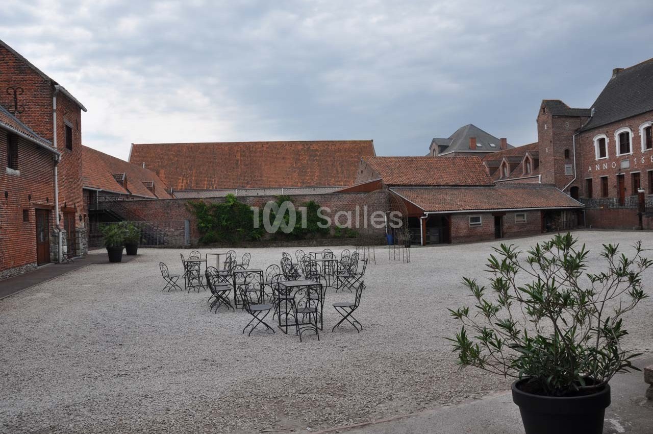 Cour pavée avec des tables et chaises en fer forgé, entourée de bâtiments en briques sous un ciel nuageux.