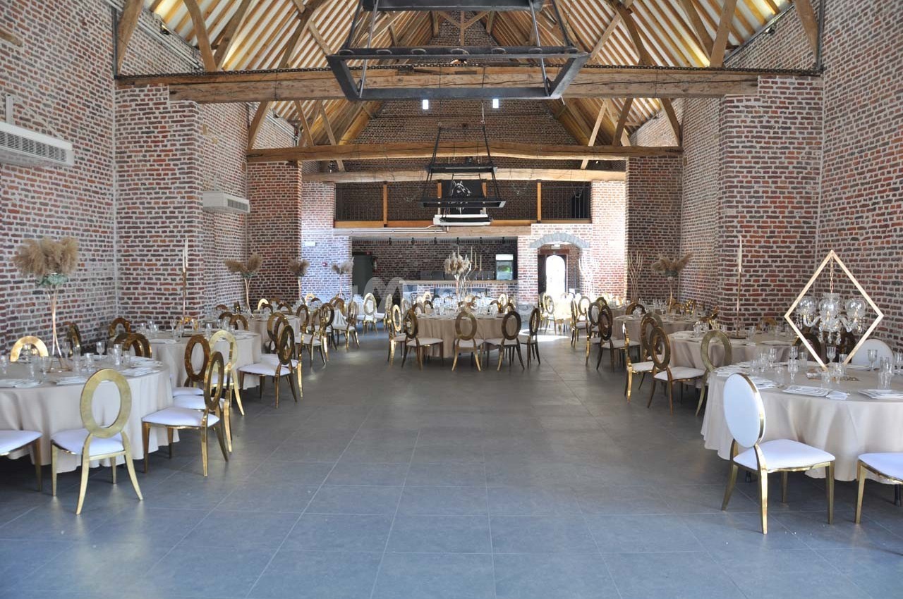 Salle de réception spacieuse avec murs en briques, poutres apparentes et tables rondes dressées pour un événement.