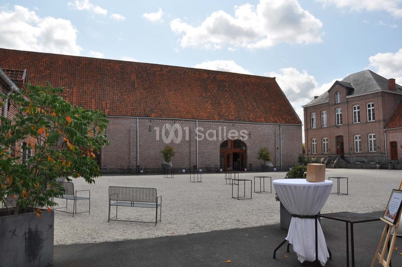 Cour pavée avec une grande bâtisse en briques et toit en tuiles, entourée de bancs et d'arbres en pot.